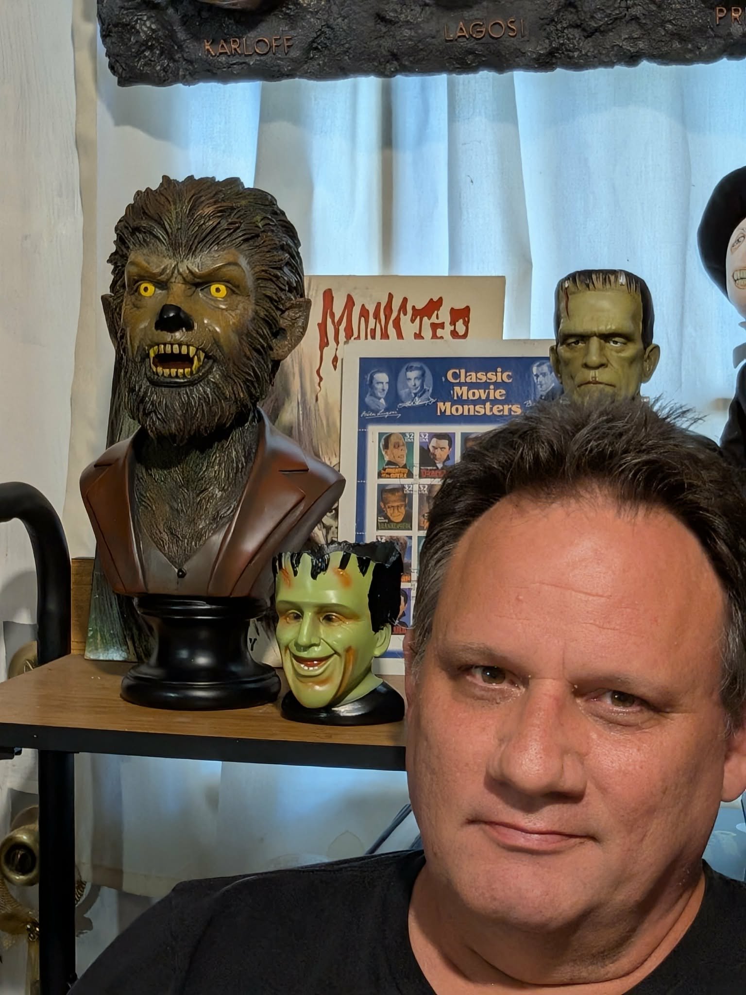 A man with short dark hair taking a selfie with Halloween or movie monster masks on display behind him. The masks include a werewolf, Frankenstein's monster, and a clown, with a poster in the background titled 'Classic Movie Monsters'.