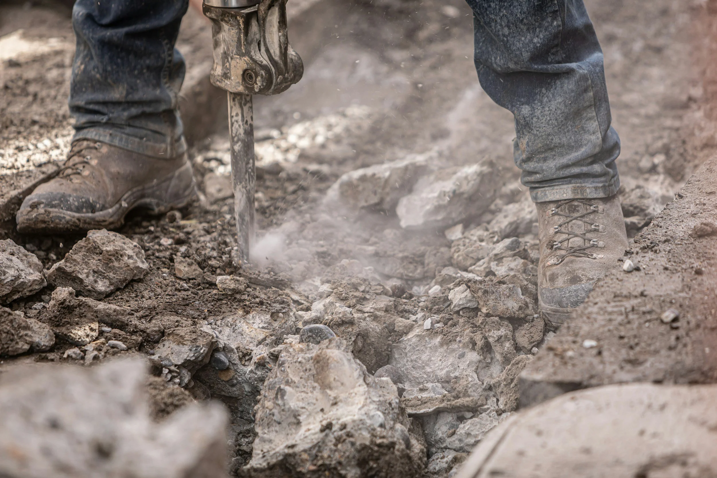 A person wearing gray work boots and gray jeans is using a jackhammer to break up concrete or asphalt on a construction site. The ground is rough and dusty with scattered debris.