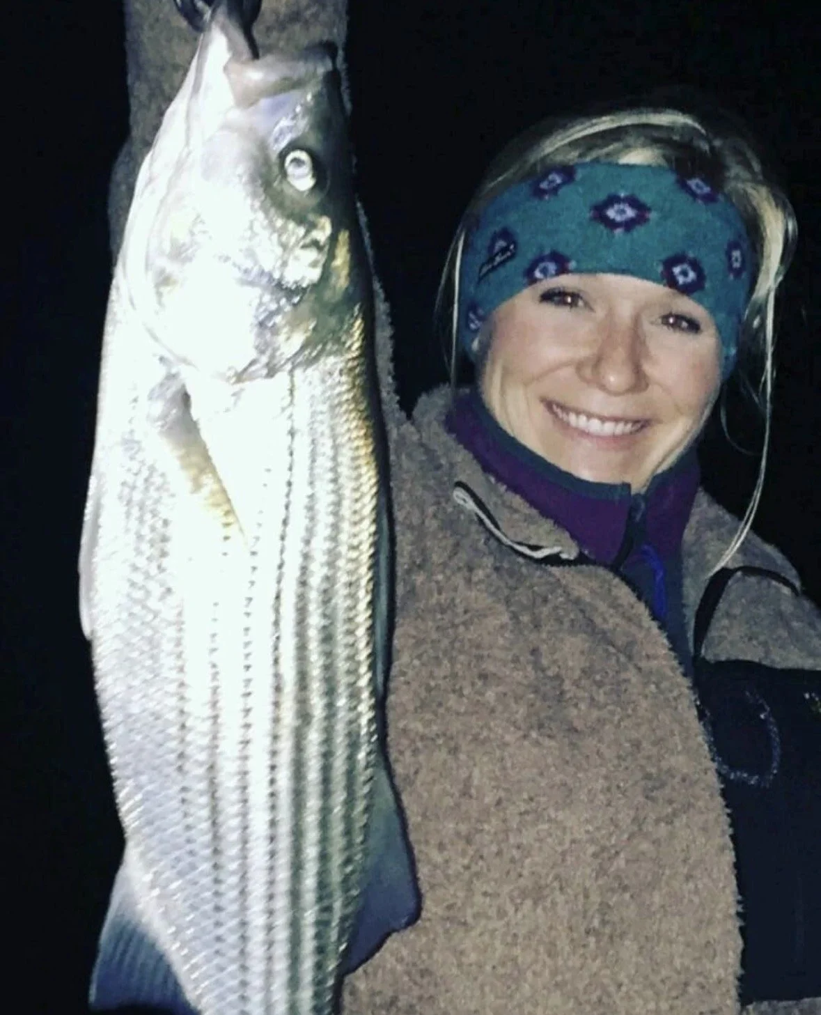 A woman smiling while holding a fish she caught, wearing a blue patterned head covering and outdoor jacket, on a dark night or in low light conditions.