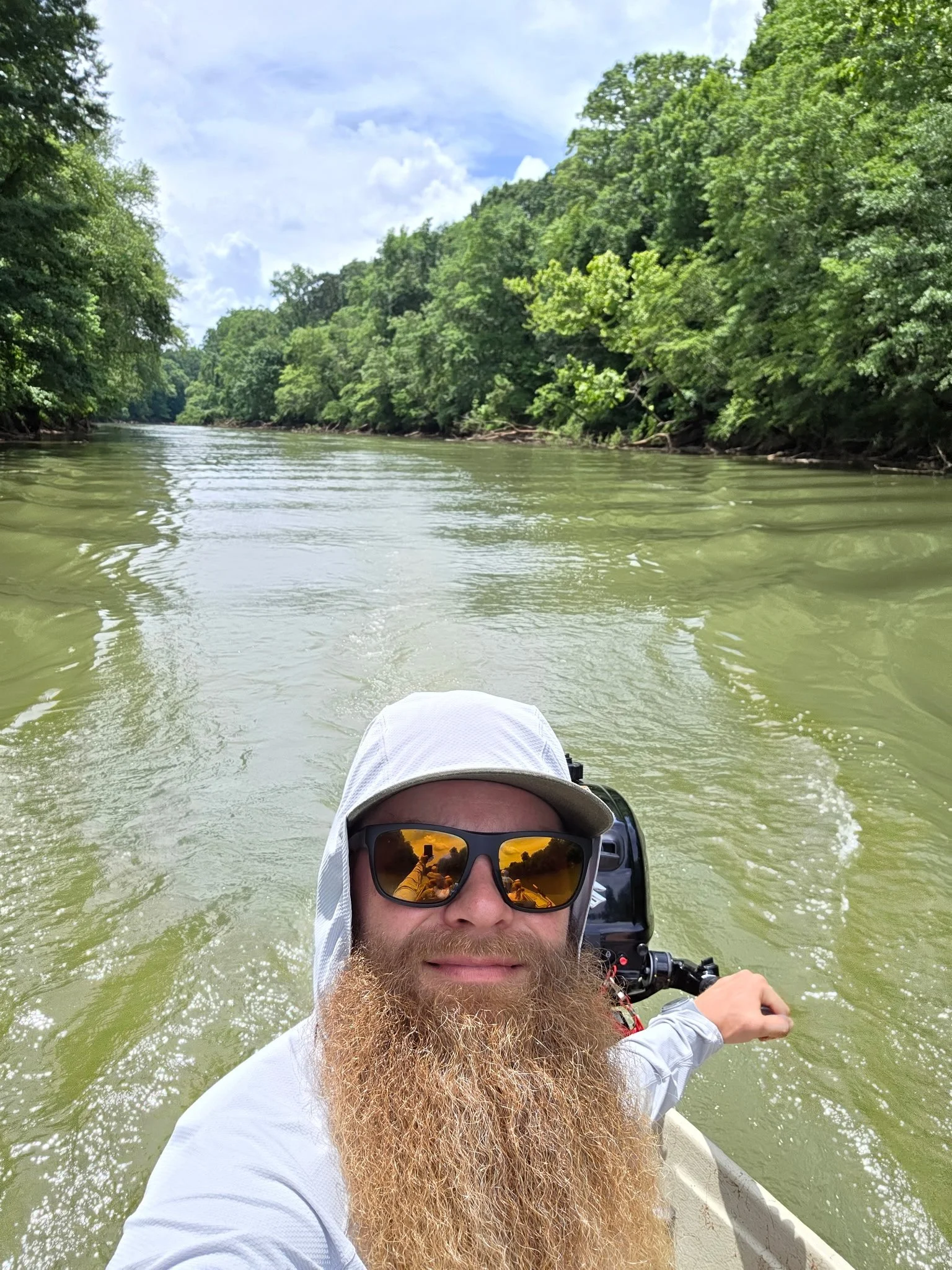 A person with a beard wearing sunglasses, a white hat with a neck cover, and a gray shirt is taking a selfie on a boat while water and lush green trees are in the background.
