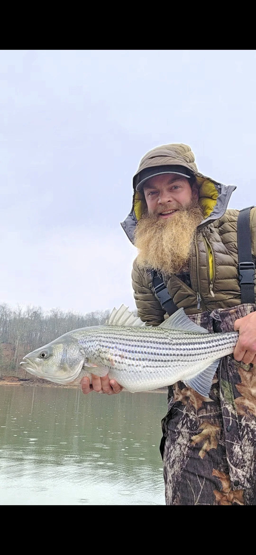 A man with a long beard and outdoor clothing holding a large striped fish near a body of water, with trees in the background under a cloudy sky.