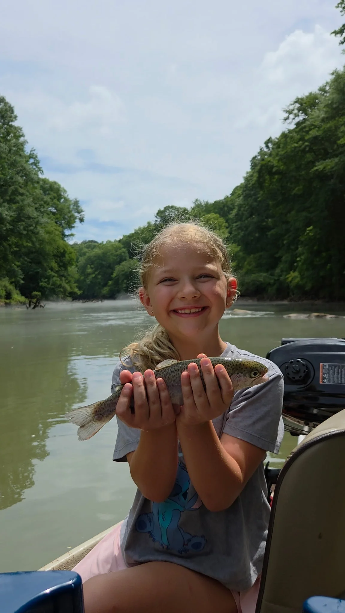 A young girl with blonde hair holding a fish on a boat in a river, with green trees and a cloudy sky in the background.