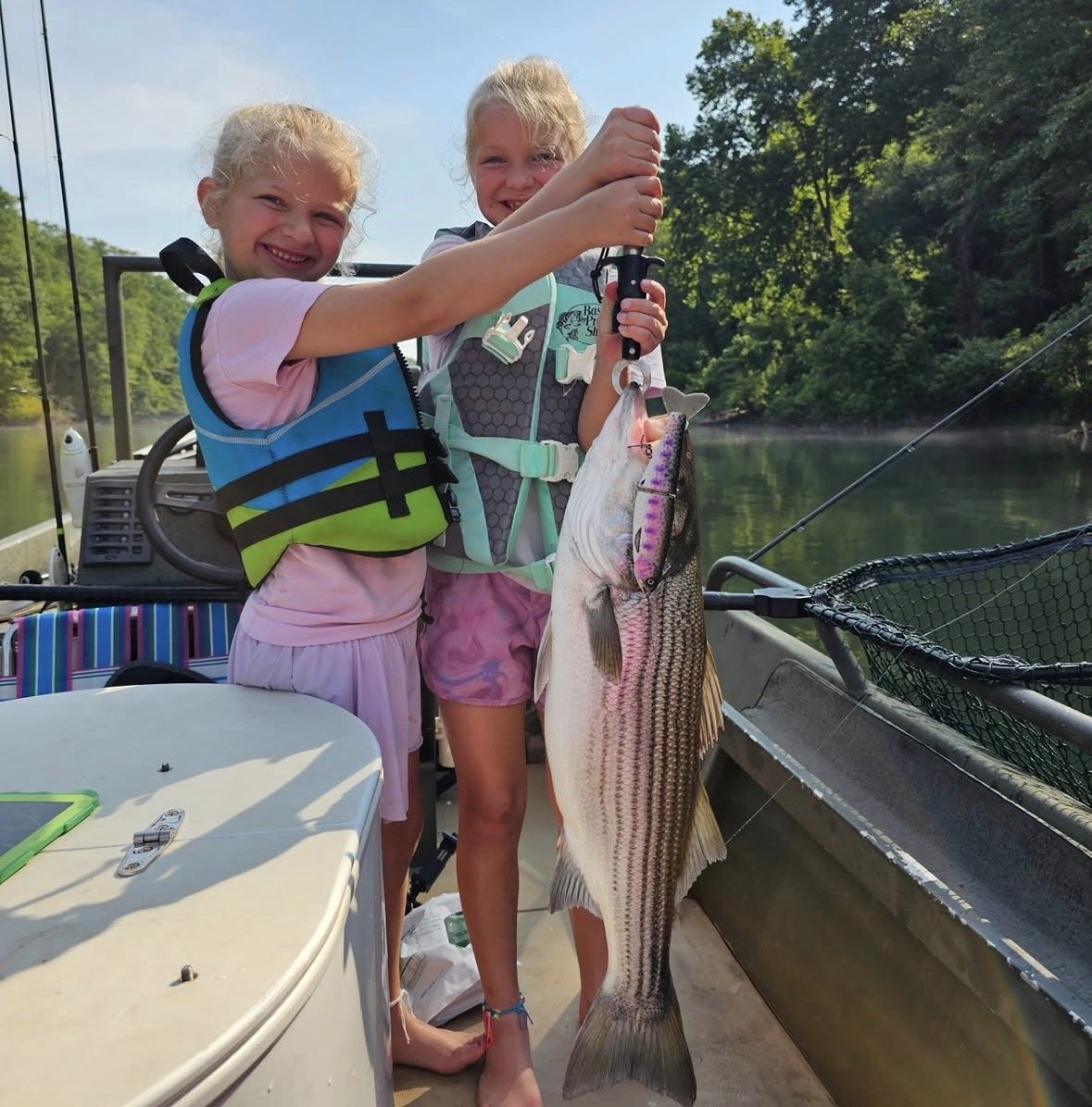 Two young girls on a boat smiling and holding a large fish they caught, with fishing gear and water in the background.