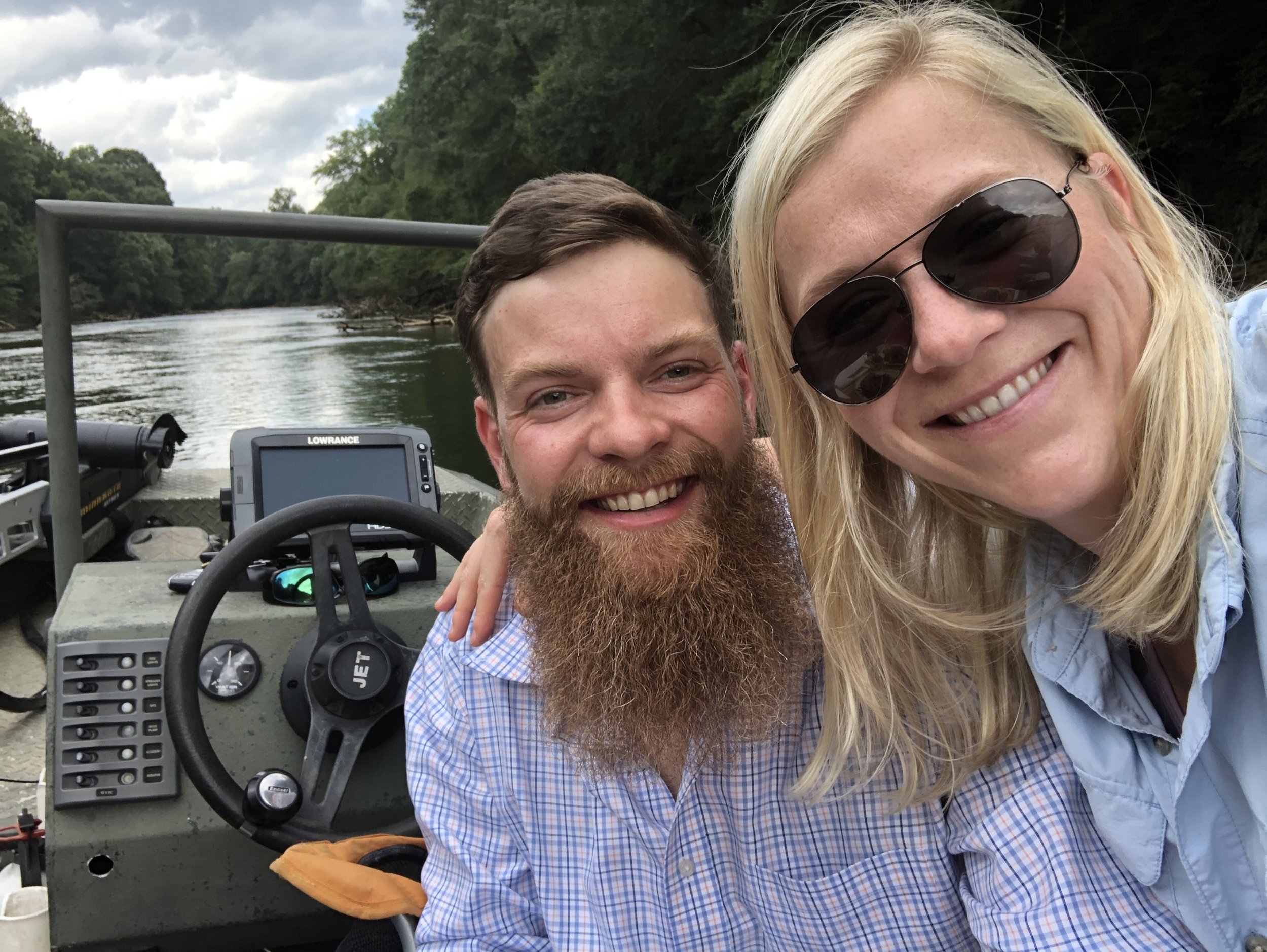 Two smiling people, a man with a full beard and a woman with blonde hair and sunglasses, taking a selfie on a boat in a river.