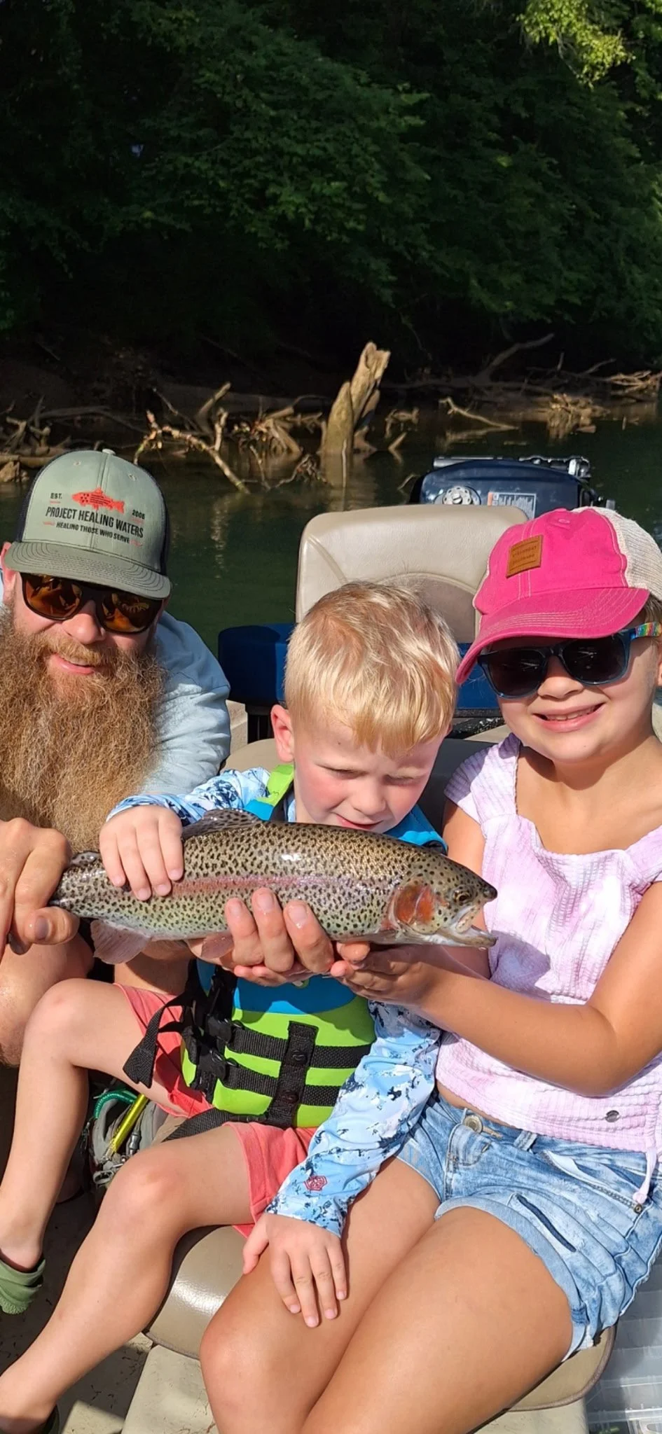 A family holding a large rainbow trout fish while sitting in a boat on a lake surrounded by trees.