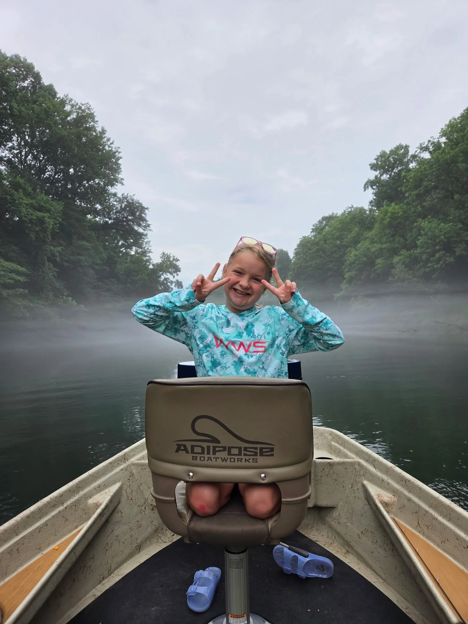 A smiling girl with blonde hair making peace signs with her hands, sitting in a boat on a calm river surrounded by green trees, wearing a blue patterned jacket, with a pair of glasses on her head. The boat has a beige seat with the logo 'Adipose Boatworks', and there are blue sandals on the floor of the boat, with water mist in the air.