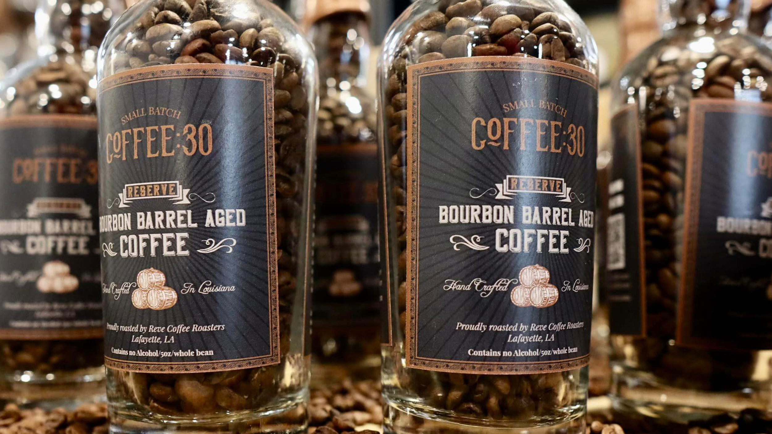 Clear plastic jars filled with roasted coffee beans on a shelf. The labels read 'Small Batch Coffee 30 Reserve Bourbon Barrel Aged Coffee,' indicating artisanal, bourbon barrel-aged coffee from Lafayette, Louisiana.