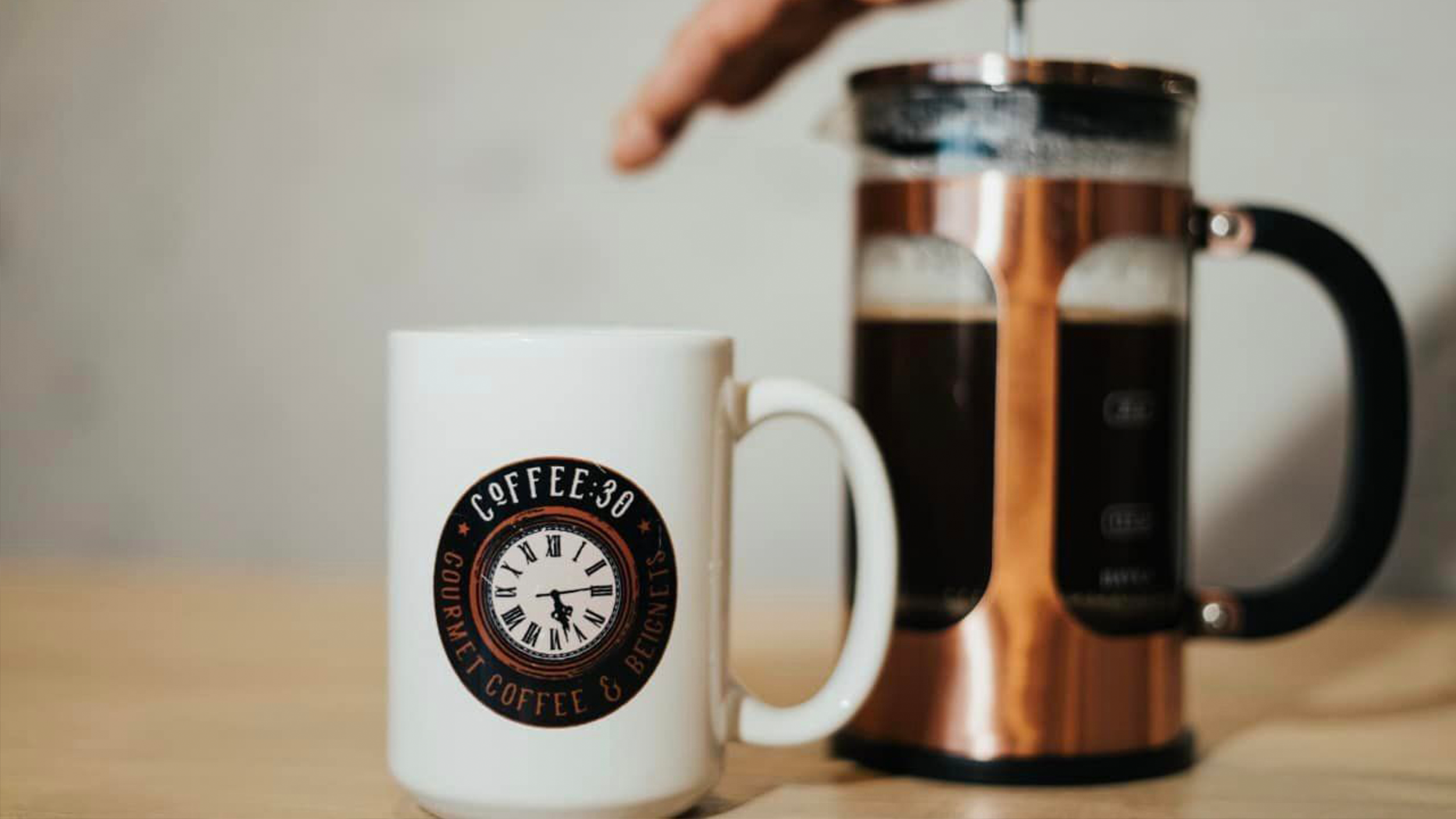 A white coffee mug with a round logo featuring a clock, a figure running, and the words 'COFFEE 30' and 'BEAUTIFUL'. Next to it, a French press with black and copper accents sits on a wooden surface.