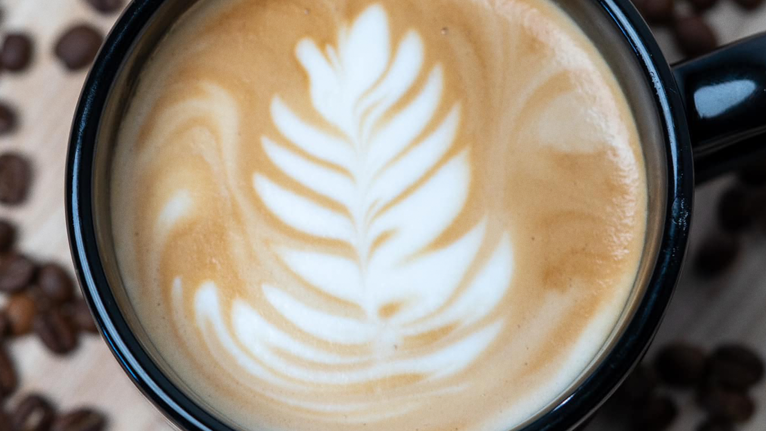 A black coffee mug filled with a latte featuring latte art in the shape of a fern or leaf, with coffee beans scattered around the mug.