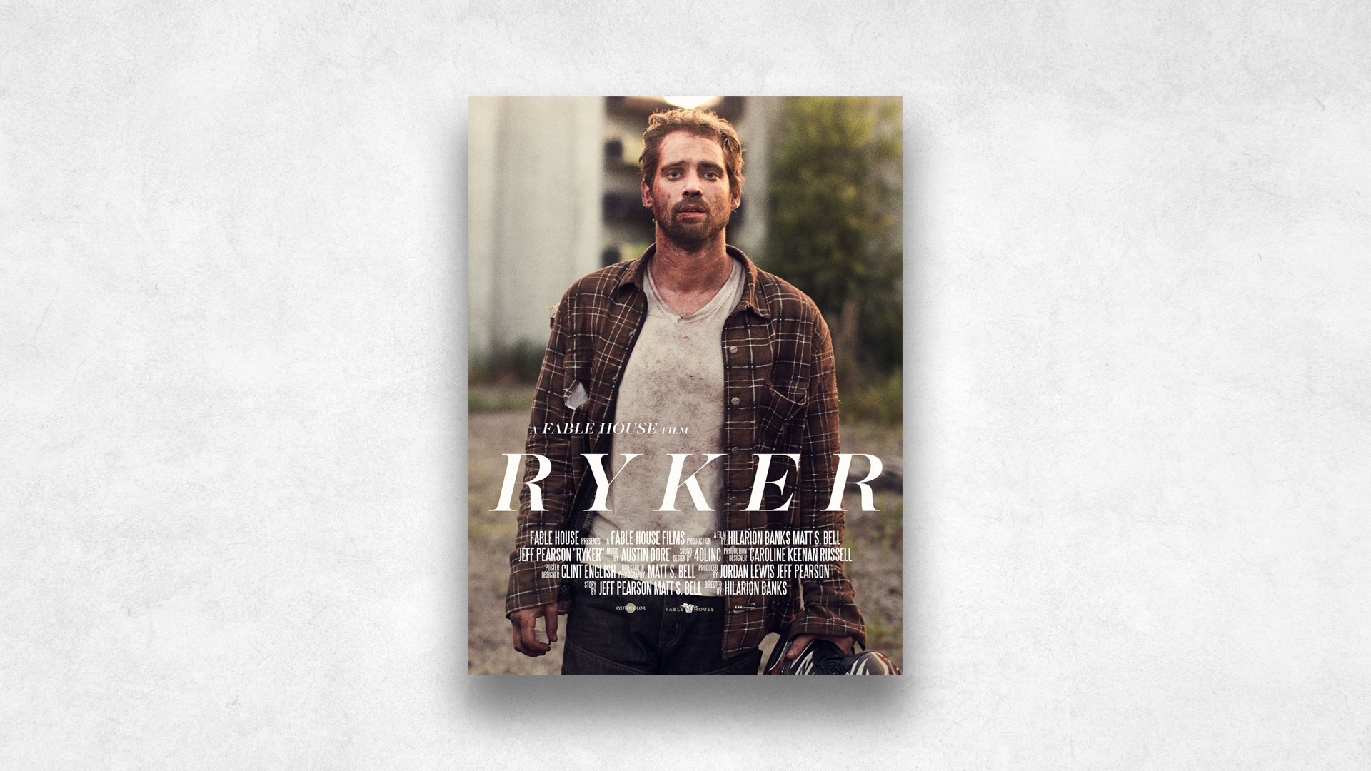 Movie poster for 'Ryker' featuring a man with brown hair, a beard, wearing a plaid shirt over a dirty white T-shirt, standing outdoors with trees and a building in the background.