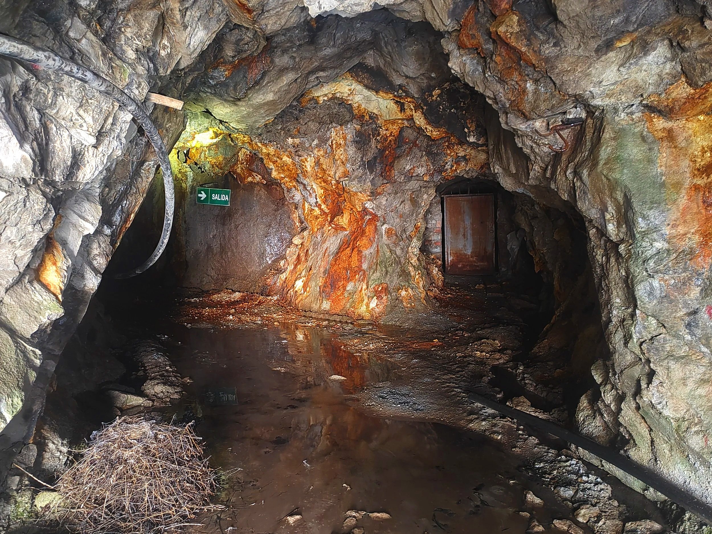 Interior de una cueva con paredes rocosas oscuras y oxidadas, con tuberías y un cartel que dice 'Salida' en la pared.