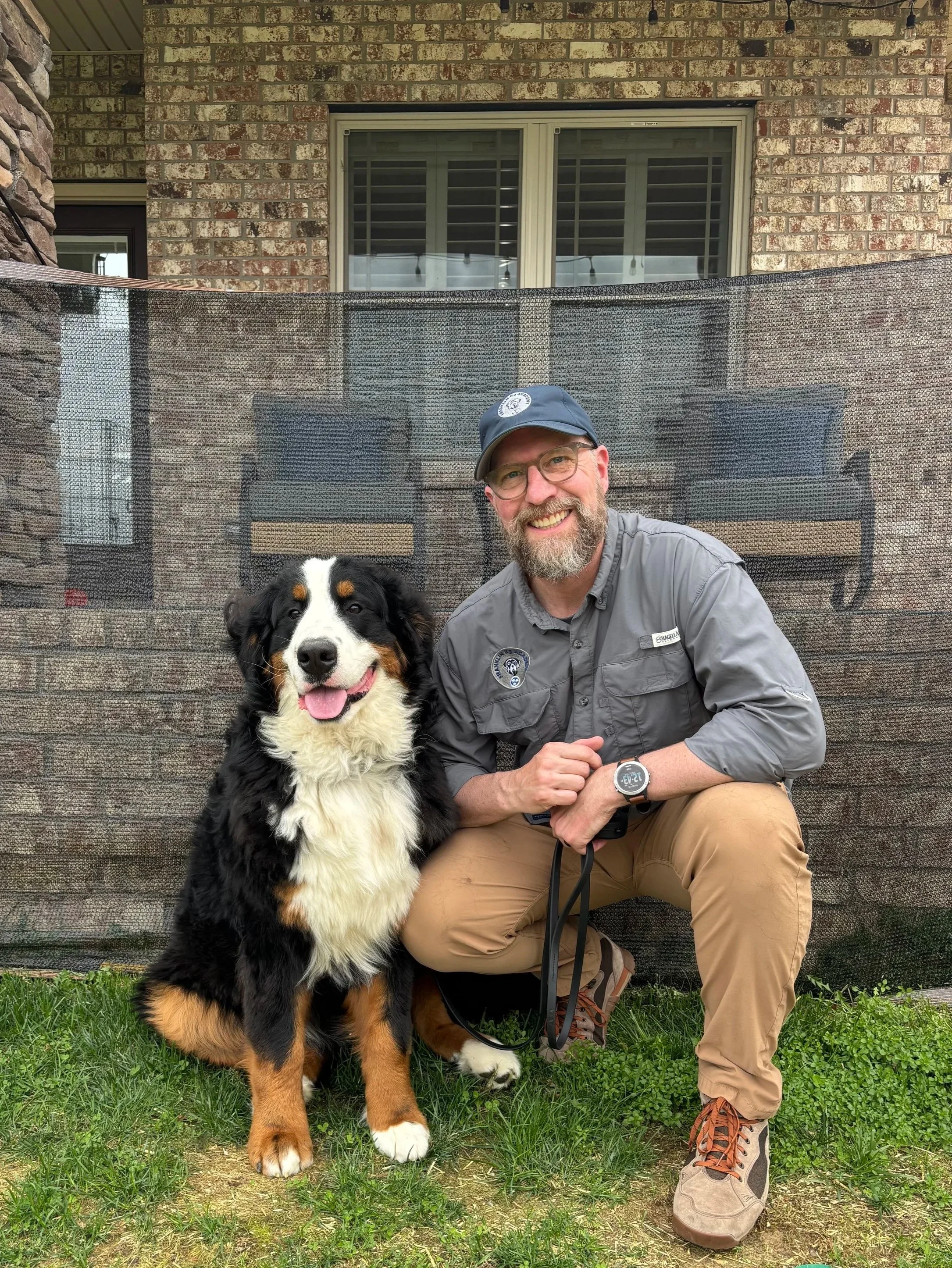 A man with glasses, beard, and a blue cap kneeling next to a Bernese Mountain Dog puppy on grass in front of a brick house with a large window. The man is smiling and holding the dog with a leash.