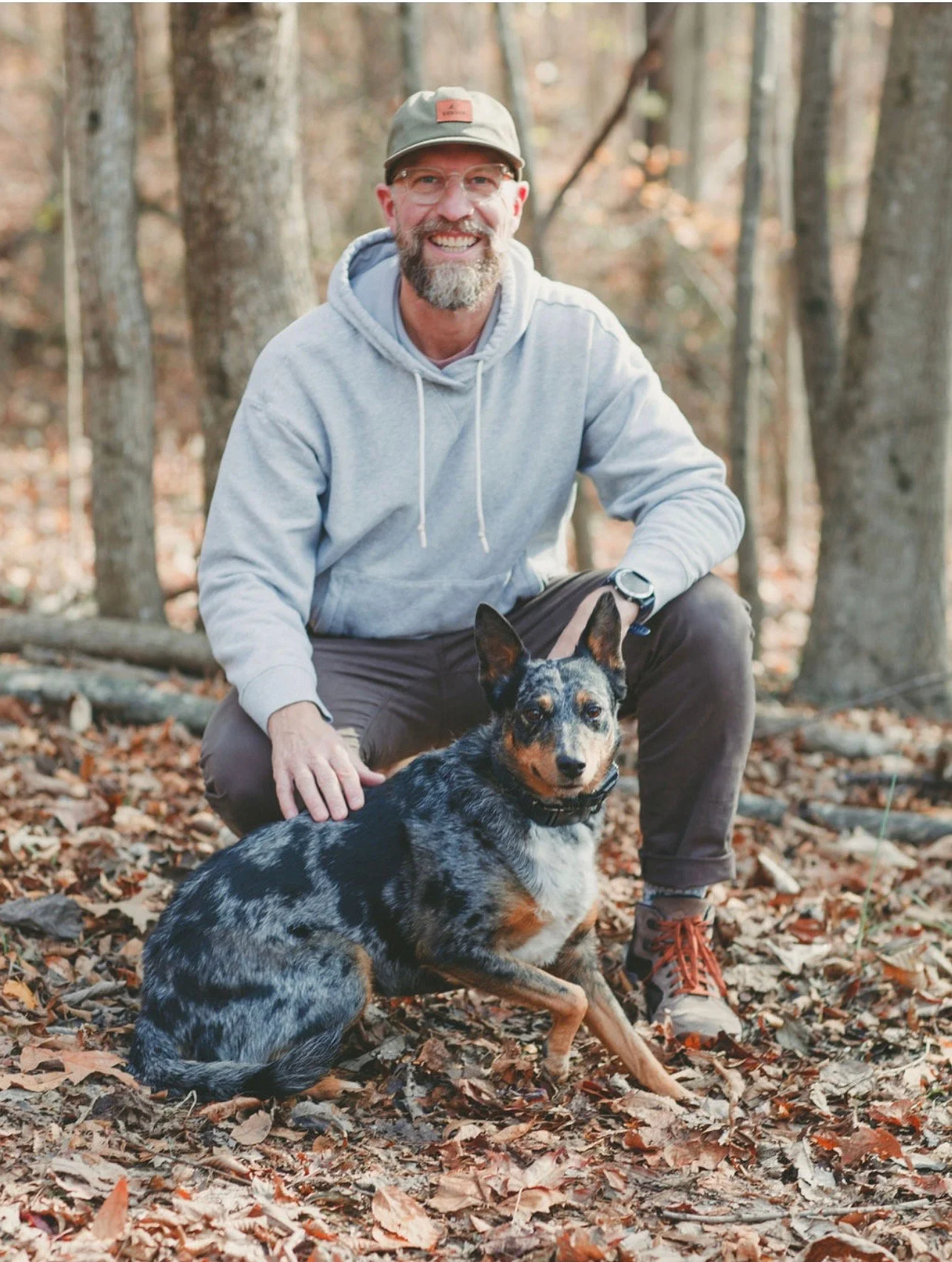 A smiling man with glasses and a bearded face in a gray hoodie, wearing a gray cap, squatting in a forest during fall, with his hand on a blue-eyed, black, gray, and tan Australian Shepherd dog sitting on fallen leaves.