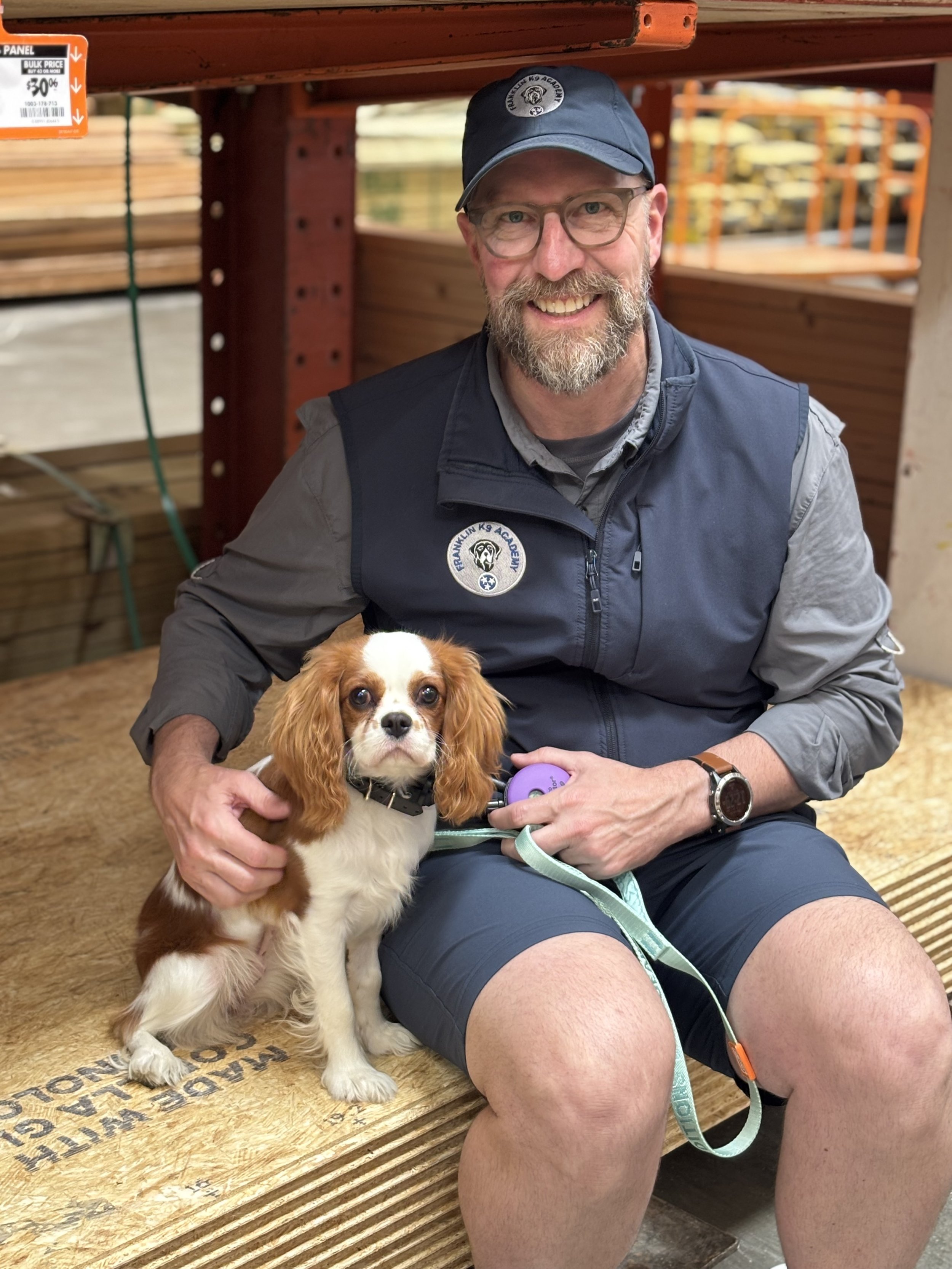 A man with glasses and a beard sitting on a wooden step, holding a brown and white dog with floppy ears. He is smiling and wearing a navy vest and cap, and a purple device is attached to his wrist. There are stacks of wood and orange metal racks in the background.