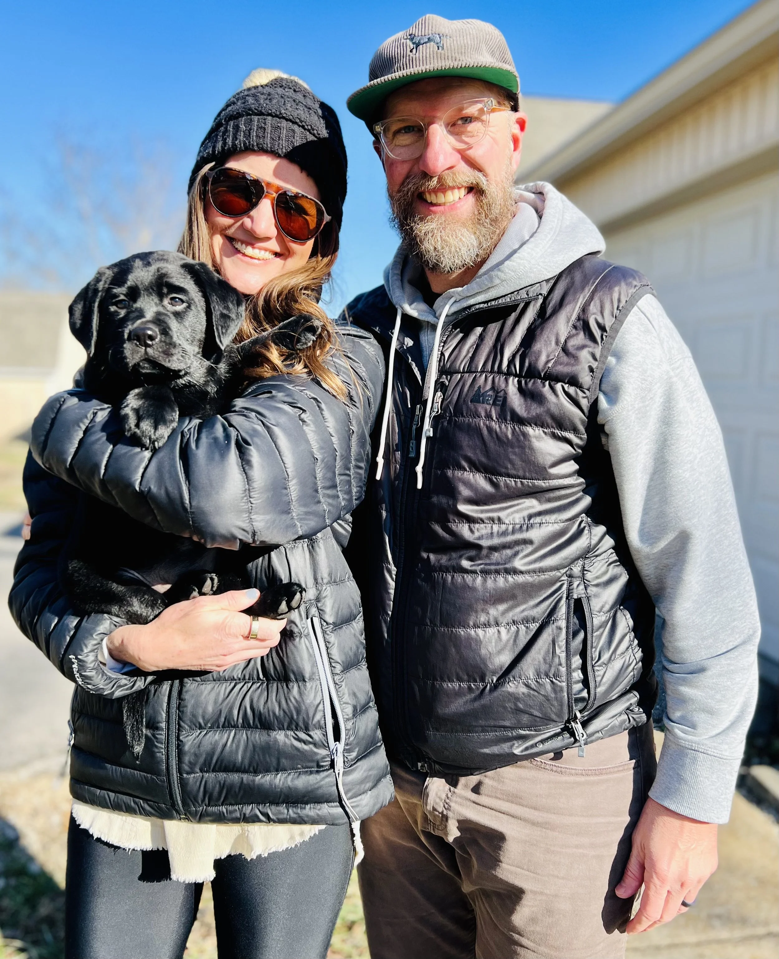 A woman and a man smiling outdoors, holding a black puppy. The woman is wearing sunglasses, a black beanie, and a puffy black jacket. The man is wearing a gray cap, glasses, and a gray hoodie with a black vest. The background shows a blue sky and a building with a sloped roof.