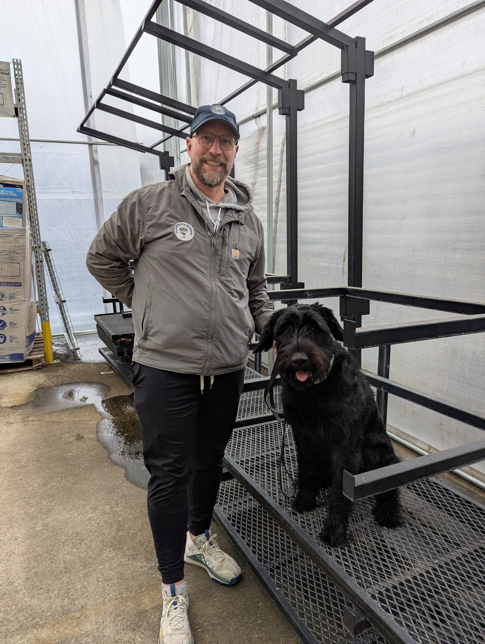 A man with glasses and a beard standing inside a greenhouse with a black dog on a leash. The man is smiling, wearing a gray jacket, a baseball cap, and white sneakers. The greenhouse has metal shelves, some plants, and a ground with water puddles.