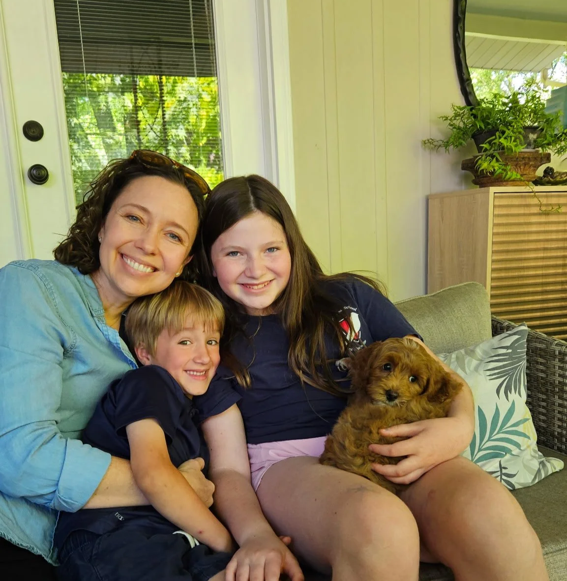 A woman and two children sitting on a couch with a small brown puppy, all smiling.