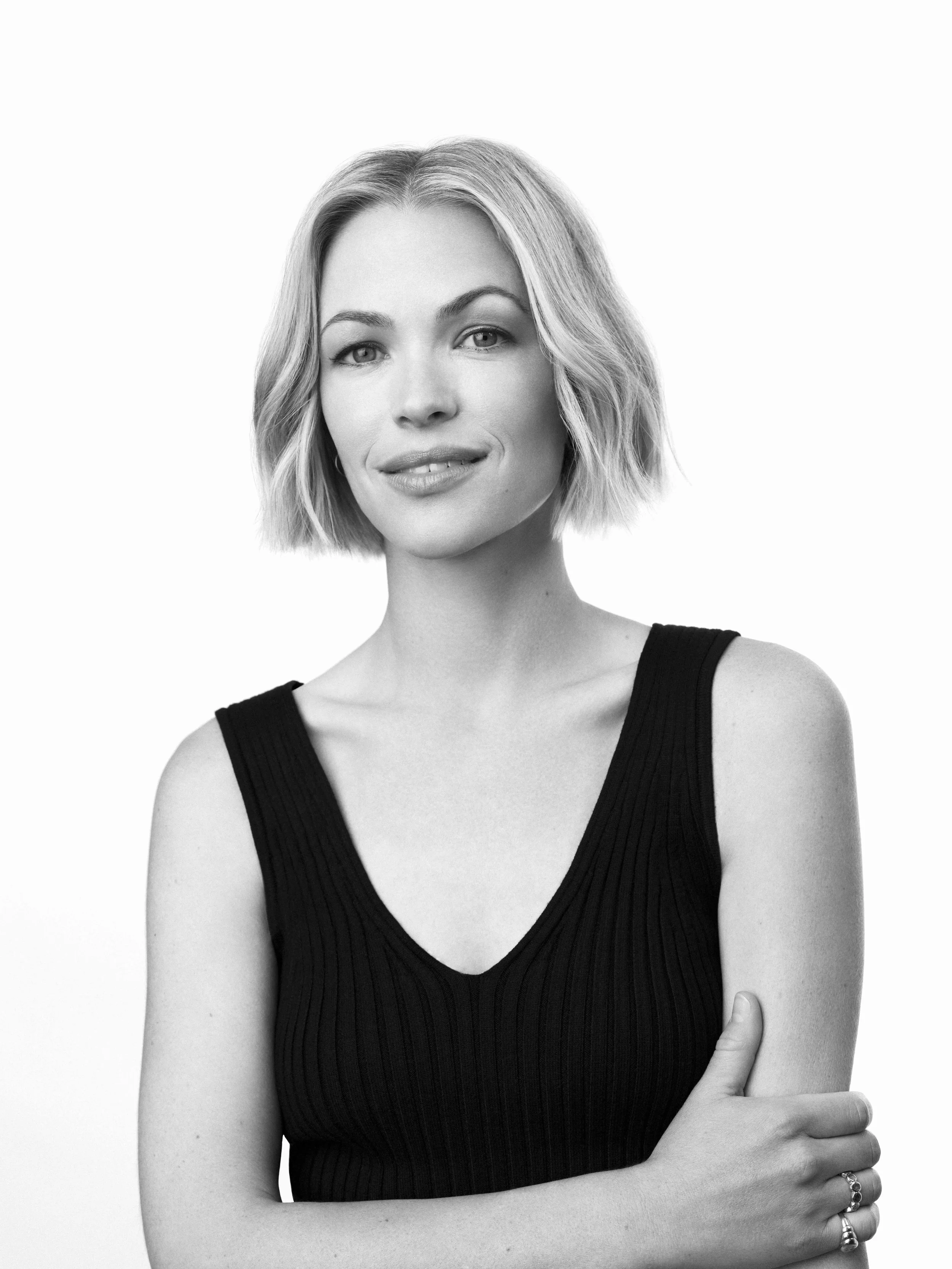 A black-and-white portrait of a woman with short, wavy hair, wearing a sleeveless black top, standing against a plain white background.