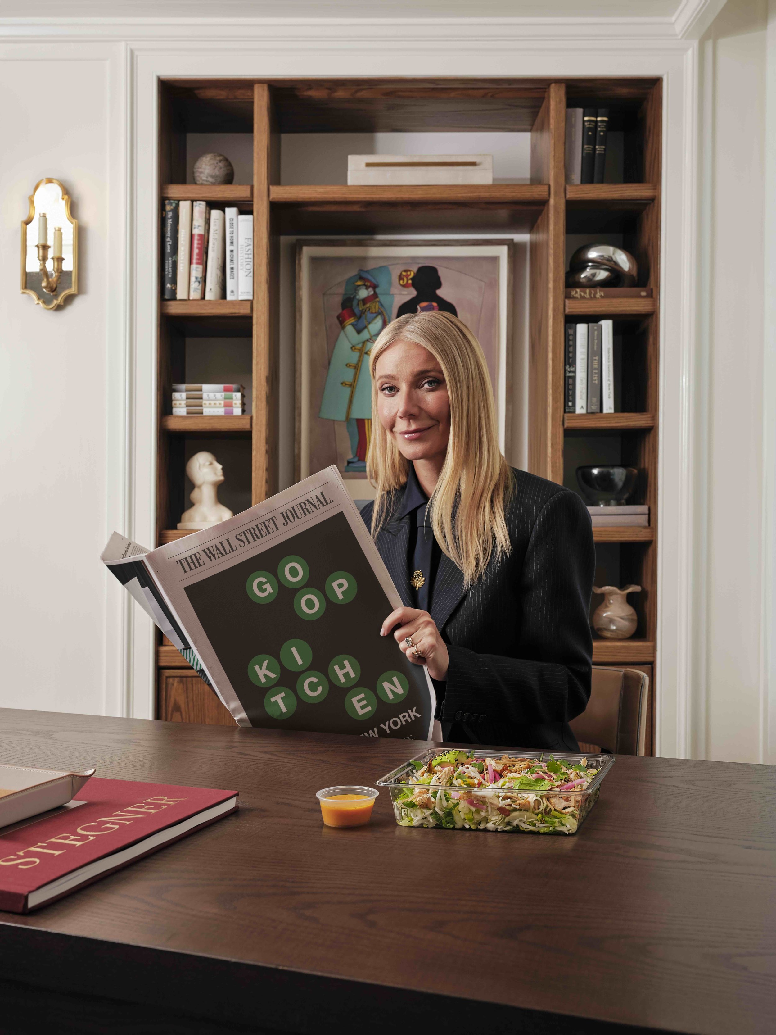 A woman with blonde hair wearing a black pinstripe suit and a brooch, sitting at a wooden table in a room, holding a newspaper labeled 'The Wall Street Journal.' On the table, there is a large salad bowl, a small cup of dressing, and a red book titled 'Designer.' In the background, there are bookshelves with various books, artwork including a framed silhouette of a woman, and decorative objects like a sculpture of a head and metallic balls.