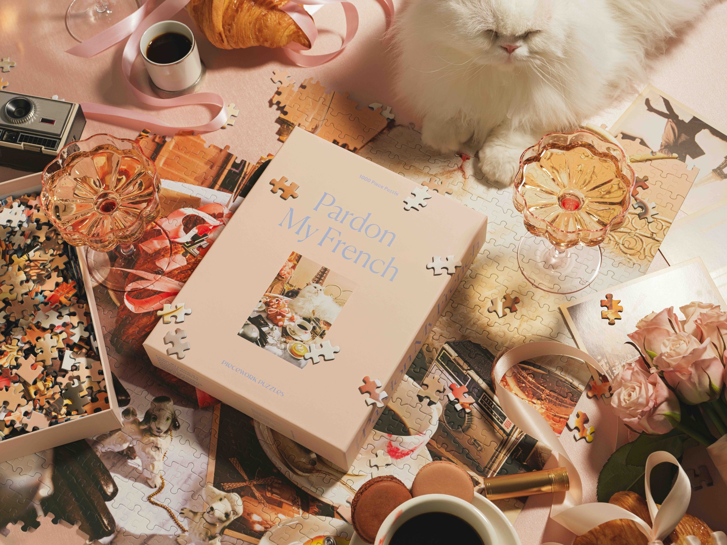 A partially completed 1000-piece puzzle featuring a romantic Paris scene, surrounded by loose puzzle pieces, two pink glass bowls, two cups of black coffee, a small vintage camera, a croissant, a box with the title "Pardon My French," flowers, a gold lipstick, and a gold necklace on a pink surface.