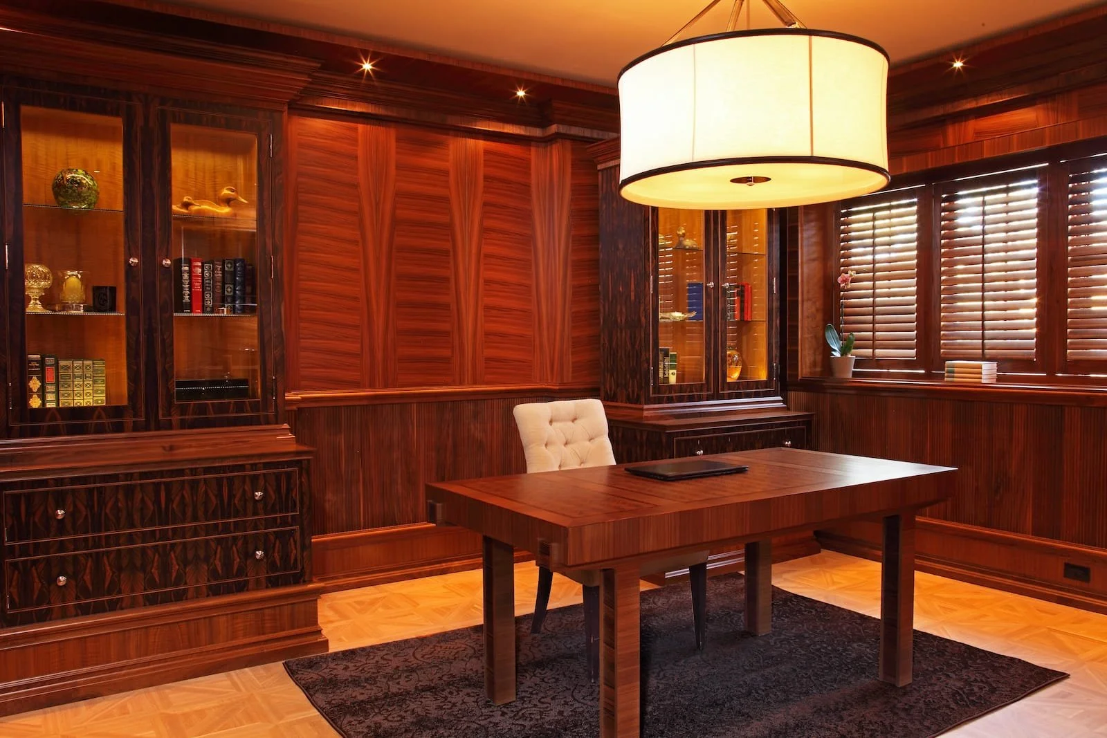 A wood-paneled office with a wooden desk and a white, tufted office chair. There are glass display cabinets with decorative items and books, and a window with wooden blinds. A large, circular ceiling light hangs above.