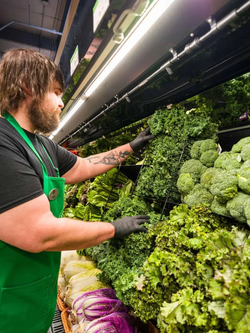 A man with a beard and tattoos wearing a green apron and black gloves working in the produce section of a grocery store.
