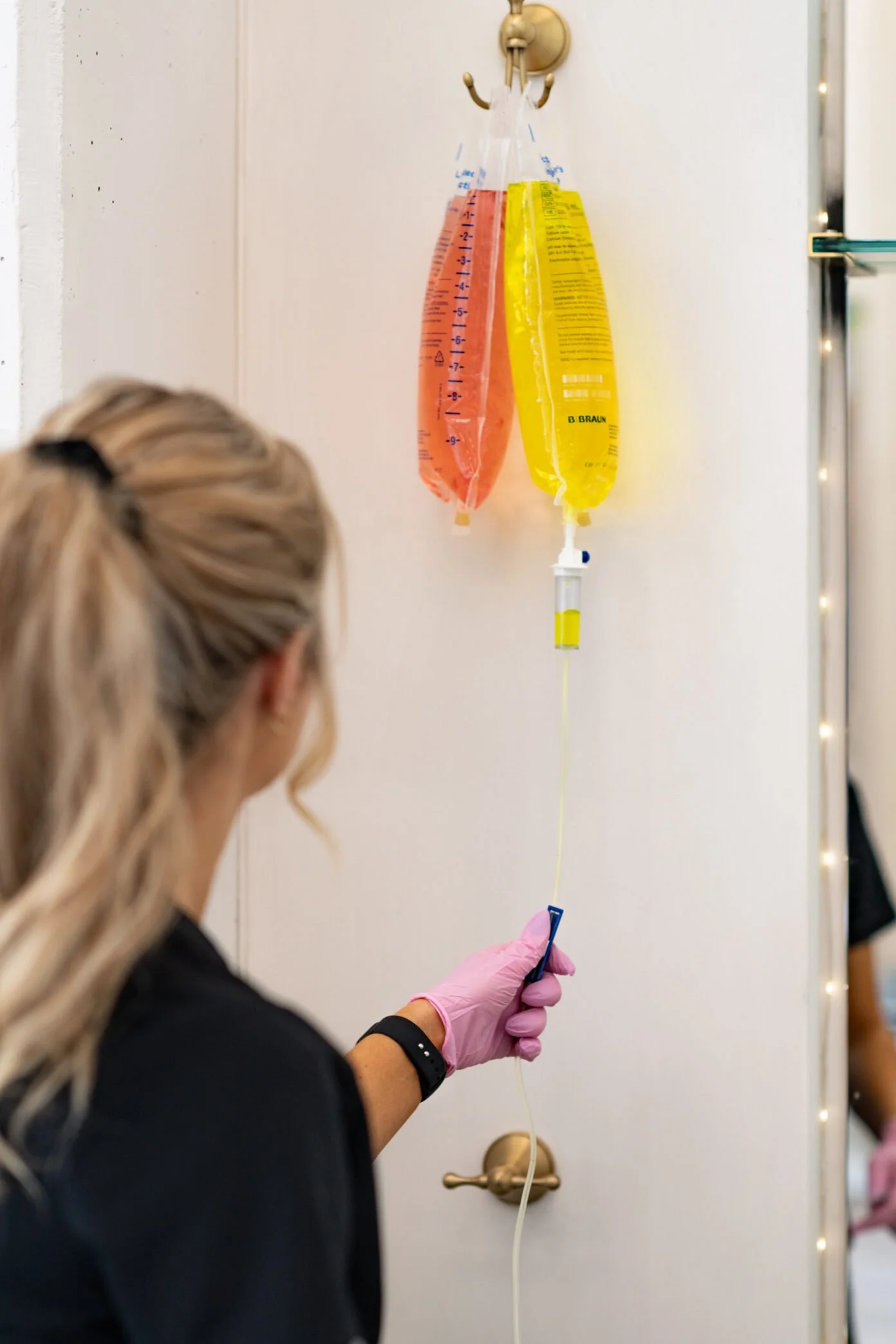 A healthcare worker in pink gloves preparing an intravenous line with a yellow medication bag hanging on a hook on a white wall.