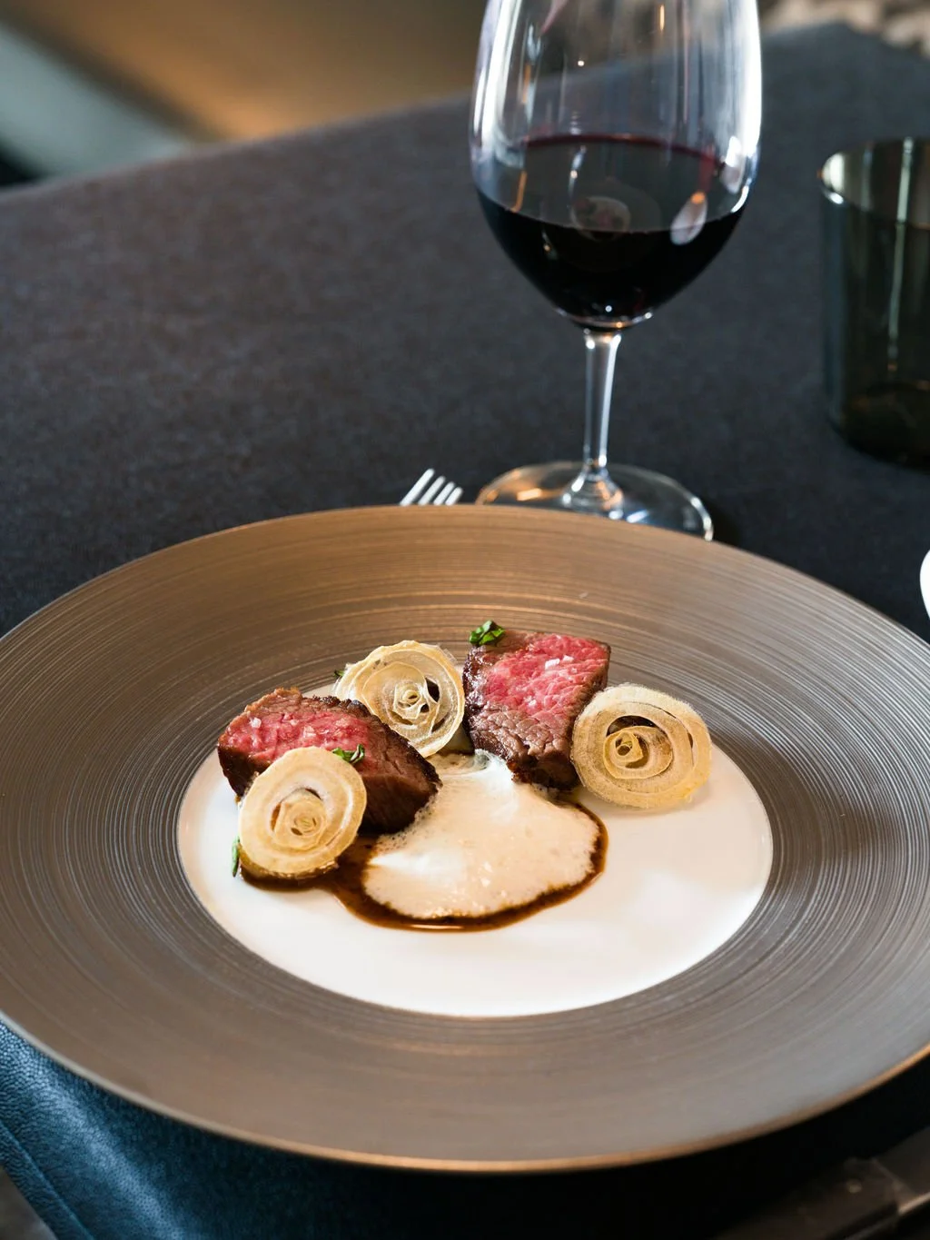 A plate with two pieces of cooked steak, garnished with sliced vegetables, served with a creamy sauce, and a glass of red wine in the background.