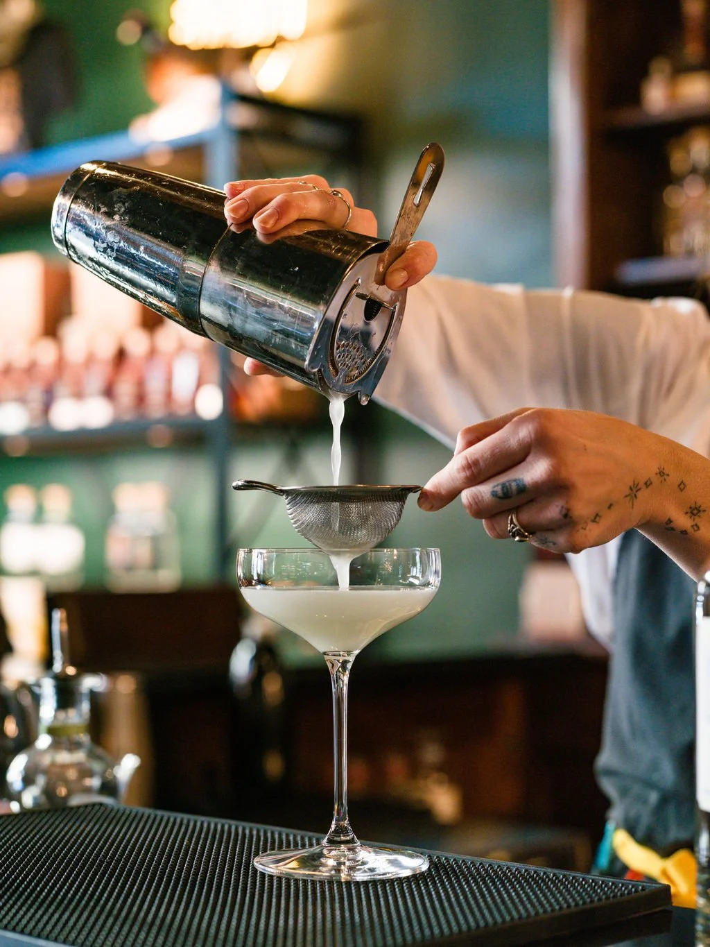 Bartender pouring a cocktail through a sieve into a martini glass in a bar setting.
