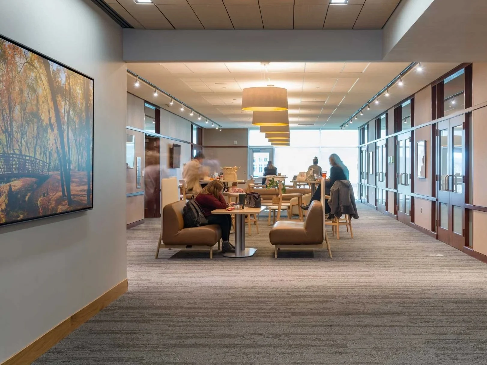 A modern office space with people sitting at tables and standing near counters, large windows at the back letting in natural light, and contemporary decor including large pendant lights and wall art.