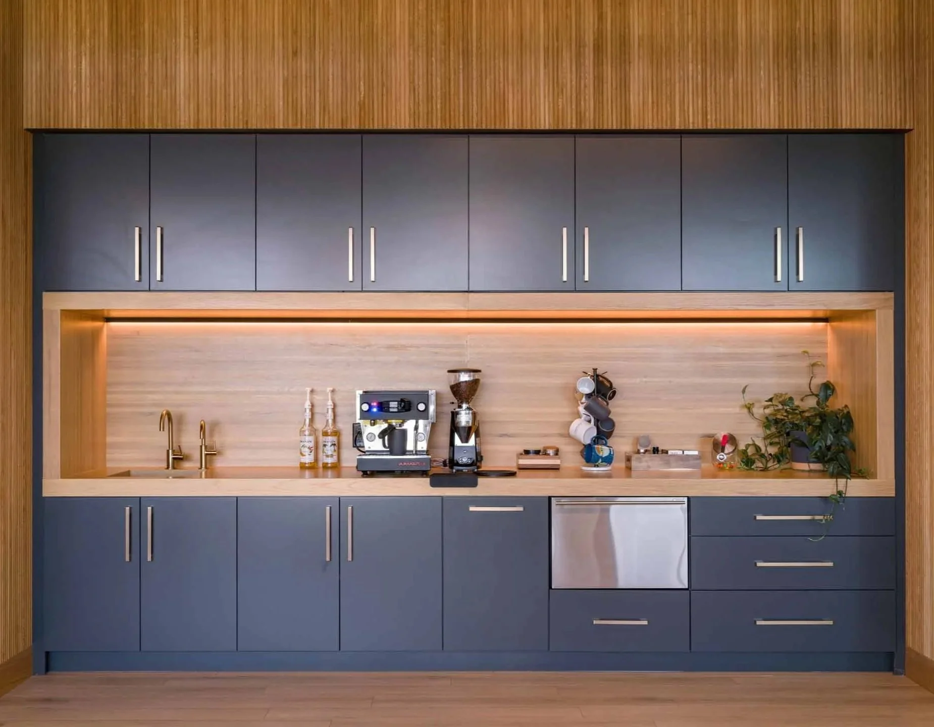 Modern kitchen with dark blue cabinets, wooden accents, and a coffee machine on the countertop, decorated with plants and mugs.