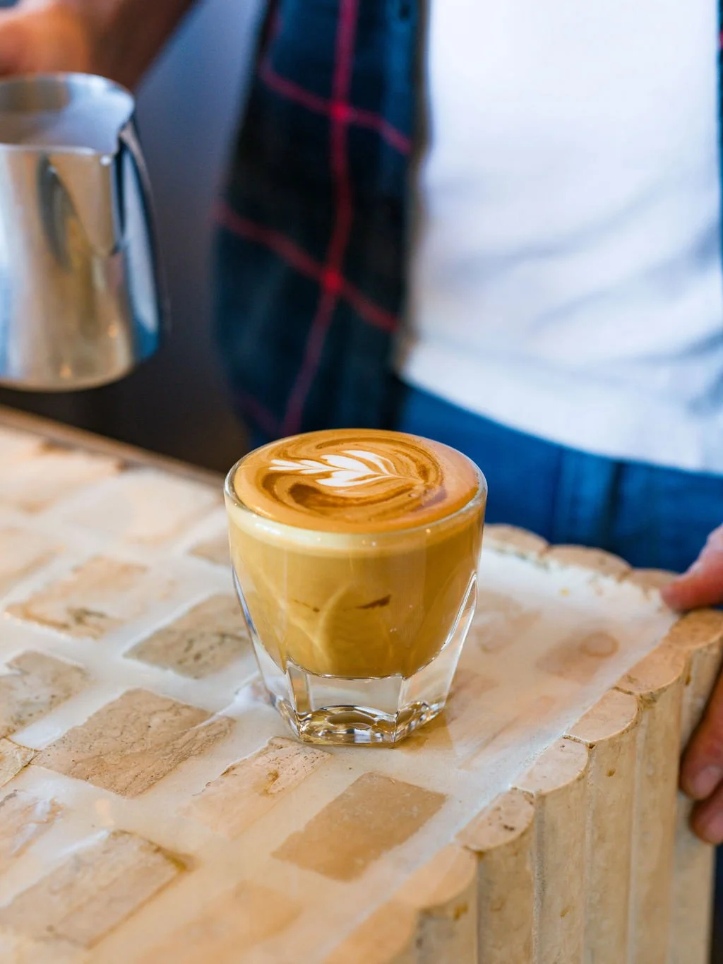A glass of coffee with latte art on top, placed on a brick countertop in a cafe or coffee shop.