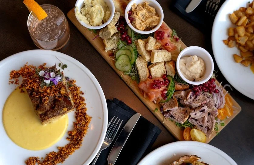 A meal featuring a plate with a piece of meat topped with breadcrumbs and a yellow sauce, a wooden board with a variety of foods including hummus, cucumber slices, bread cubes, smoked salmon, cherry tomatoes, and slices of cooked meat, and a glass of iced beverage with an orange garnish.