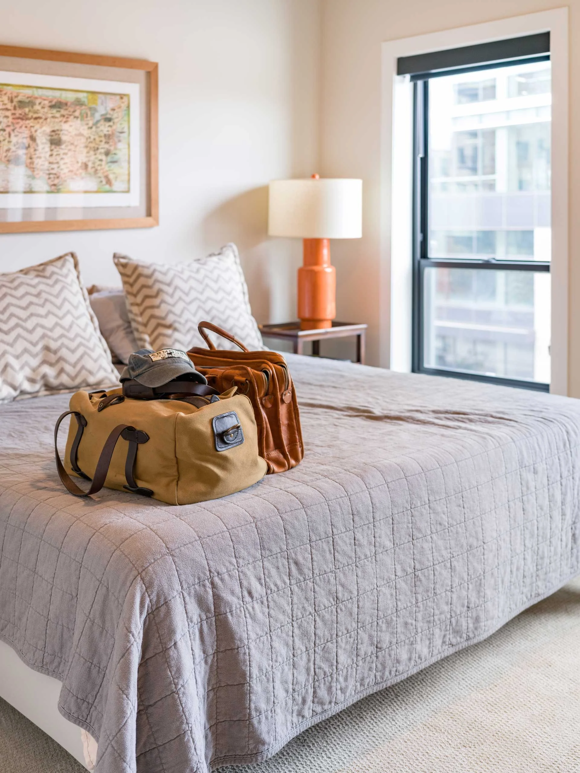 A bedroom with a bed, two pillows with a zigzag pattern, a table with a lamp, a framed map on the wall, and a window showing buildings outside. There are travel bags and a cap on the bed.