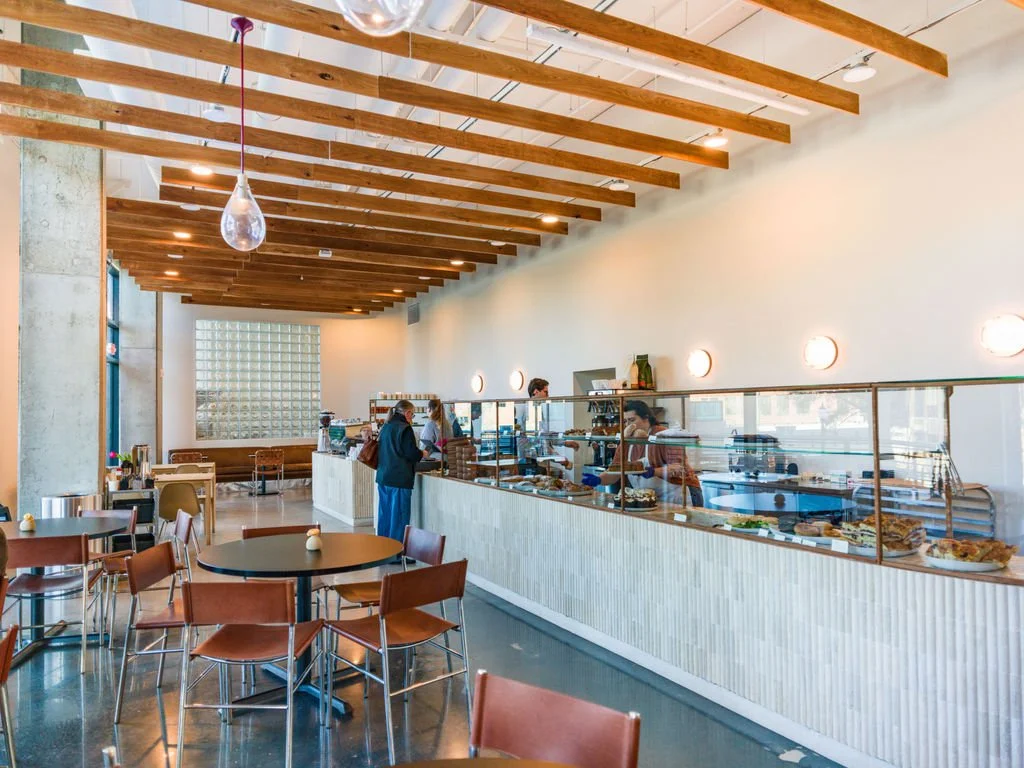 Interior of a modern cafe with a buffet counter, bar-height tables with chairs, and customers serving themselves, decorated with wooden ceiling beams and minimal lighting.