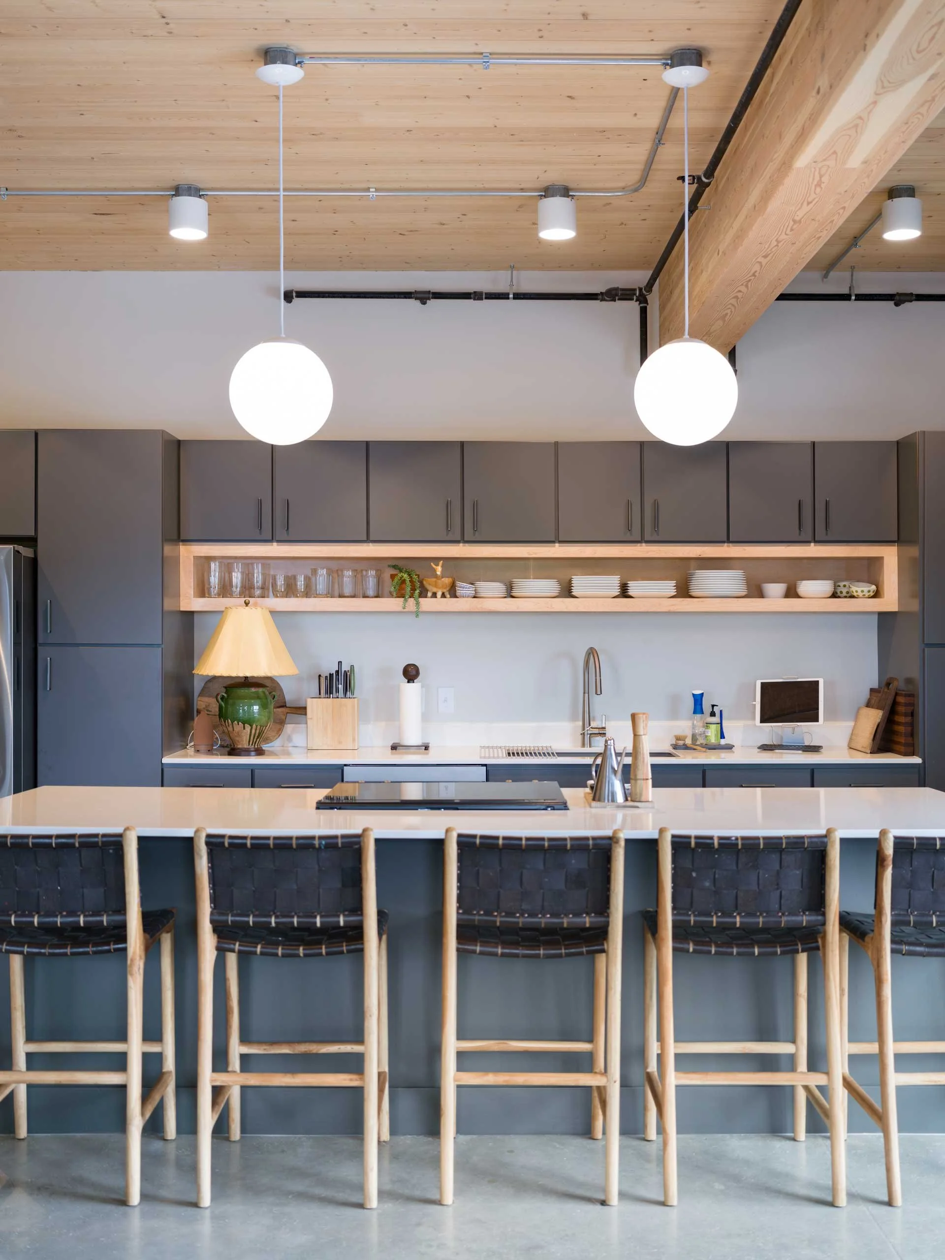 Modern kitchen with gray cabinets, open shelving with plates and bowls, a kitchen island with bar stools, and pendant lights hanging from a wooden ceiling.