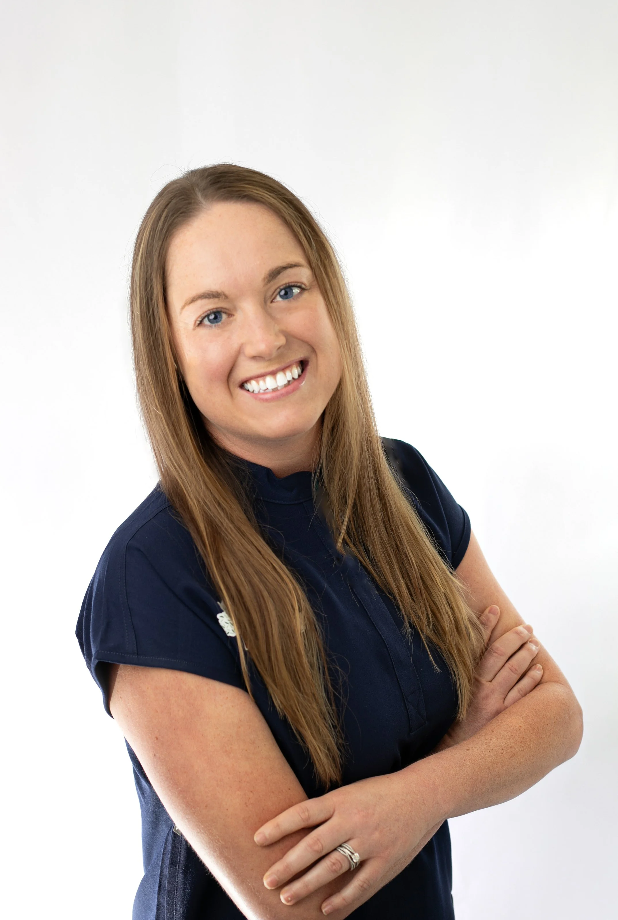 A woman with long red hair, blue eyes, and light skin, smiling and crossing her arms, wearing a navy blue shirt, standing against a plain white background.