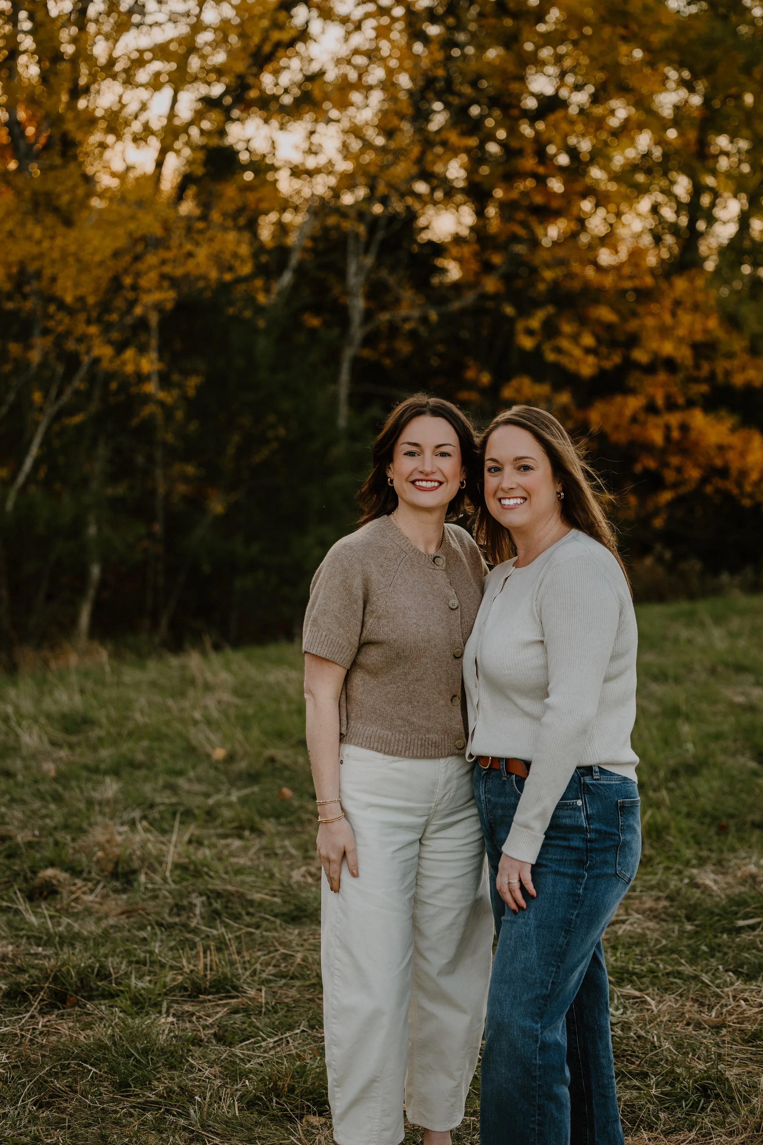 Two women standing outdoors in front of autumn trees, smiling.