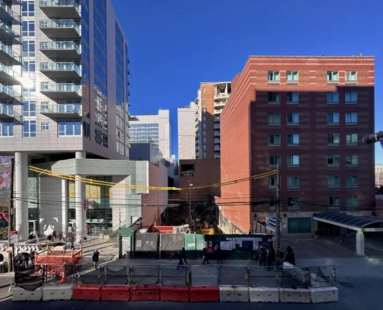Urban street scene with construction site, high-rise buildings, and pedestrians under clear blue sky.