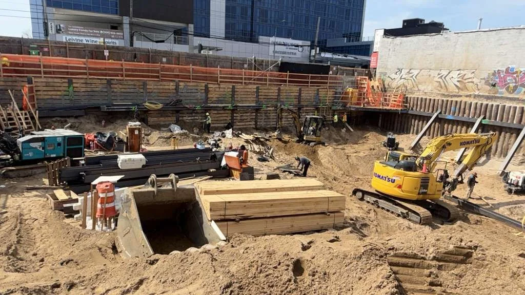 Construction site with workers, piles of wood, a small excavator, and ongoing excavation work in an urban area.