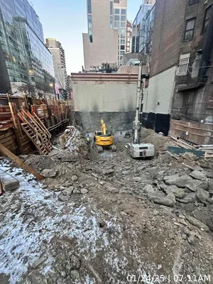 Construction site with dirt, rocks, and construction vehicles surrounded by tall buildings.