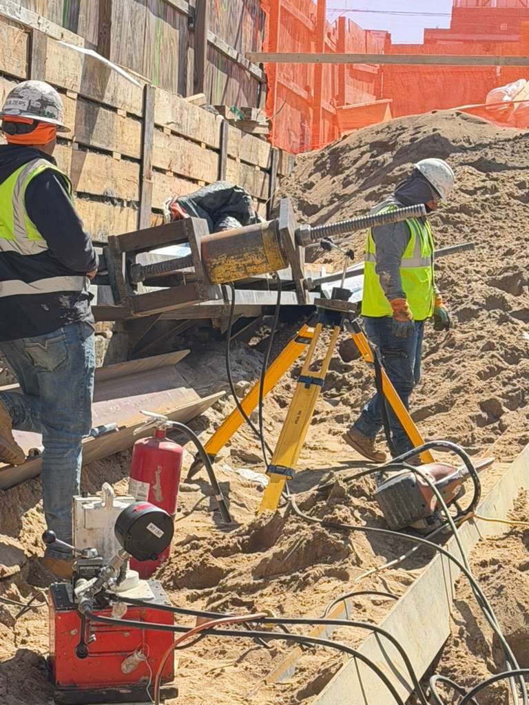 Construction workers in safety gear working on a sandy construction site with equipment including a tripod-mounted drill, fire extinguisher, and various tools. Sand dunes and wooden barriers are visible in the background.