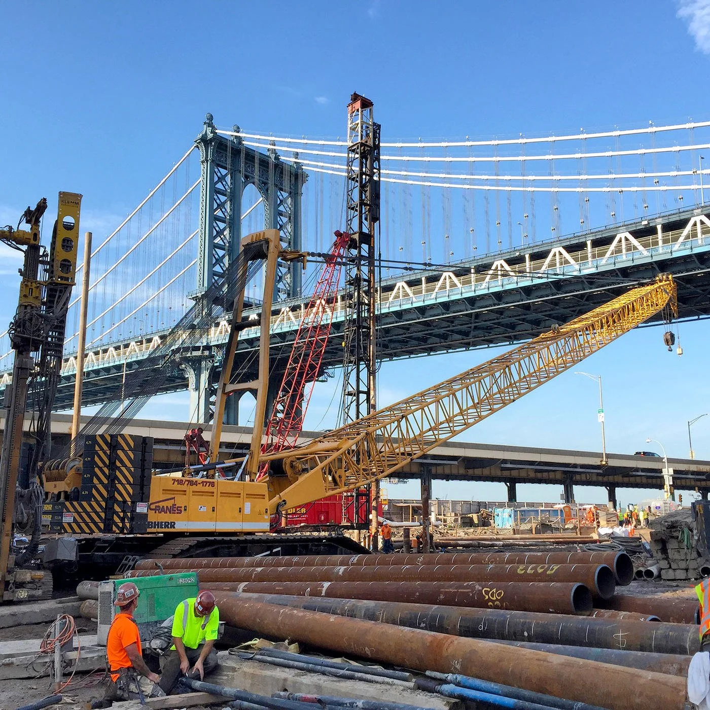 Construction workers and heavy machinery working under a bridge on a clear day.