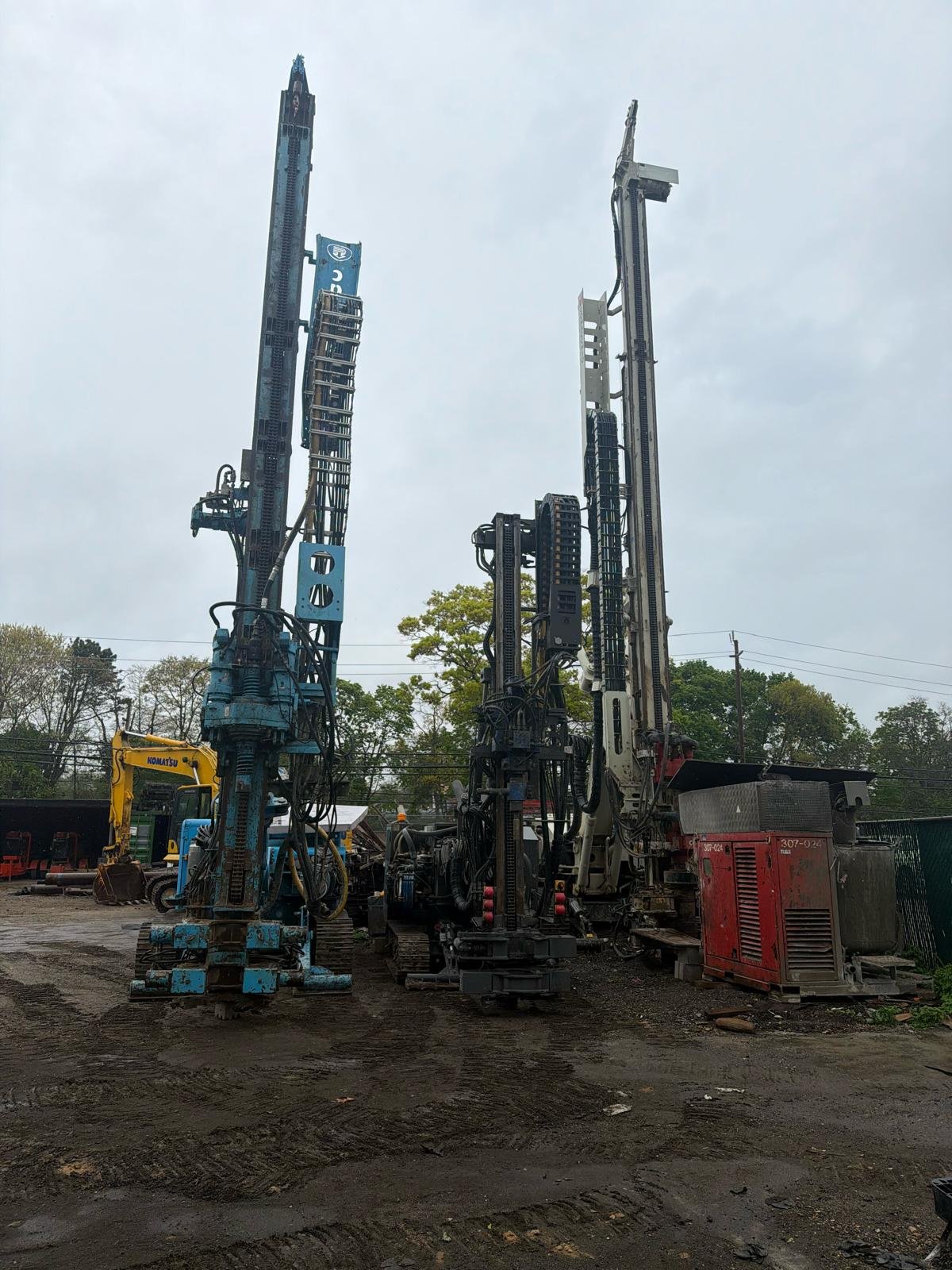 Three large drilling rigs at a construction site with trees and power lines in the background.