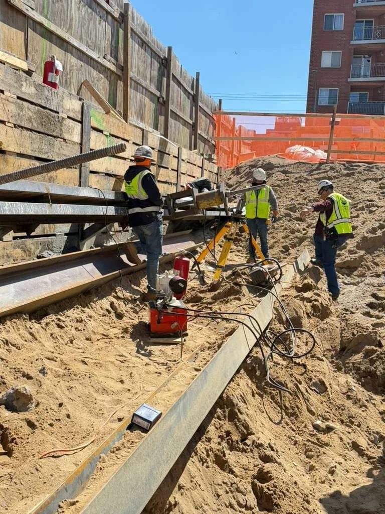 Construction workers in safety gear working on a slope at a construction site with sand, steel beams, and equipment, with a wooden retaining wall and orange safety fencing in the background.