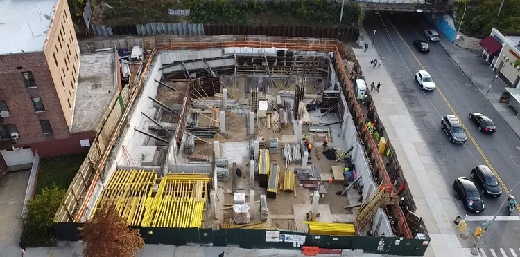 A construction site in an urban area, with concrete foundation work and yellow safety barriers, surrounded by fencing, adjacent to a street with cars and pedestrians.