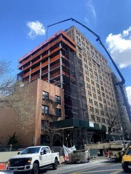 Construction site with a tall building under development, crane, and construction vehicles in front, against a blue sky.