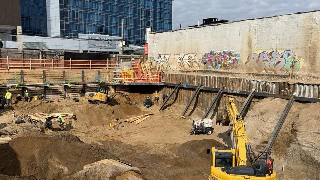 Construction site with workers, excavators, and equipment excavating soil next to a city building with graffiti on a wall.