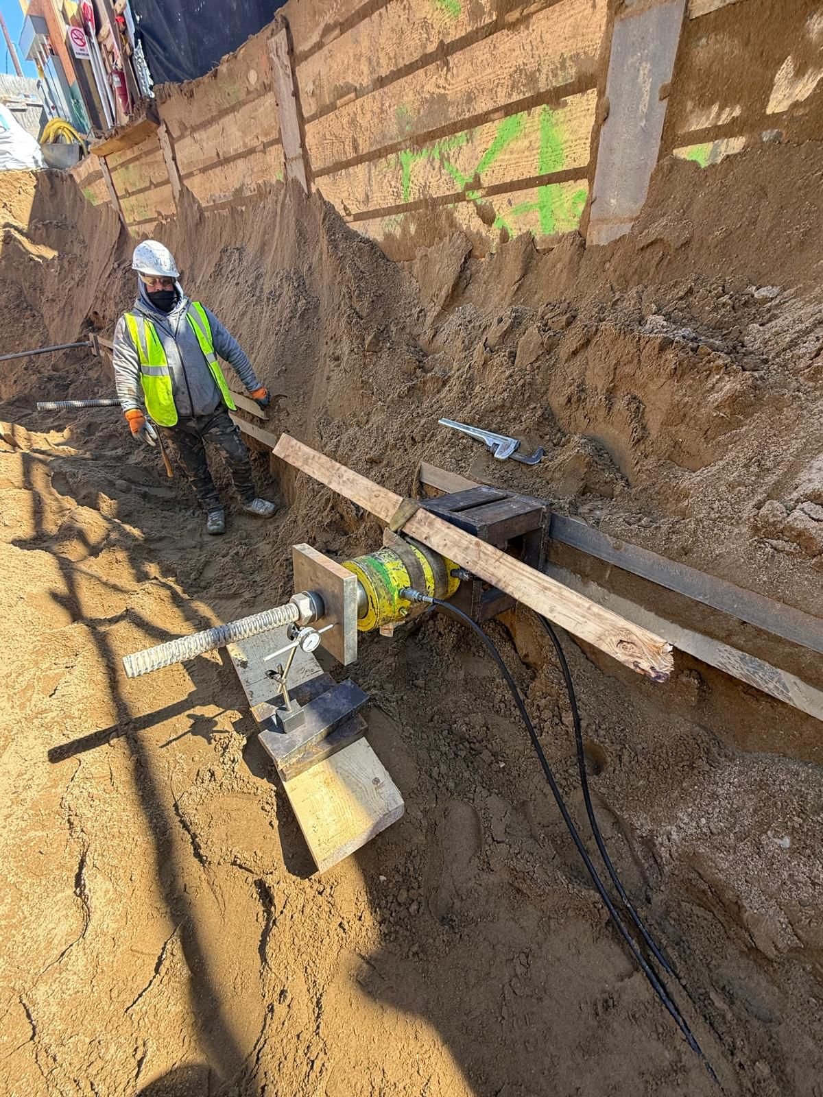 A construction worker in safety gear observes a large equipment cylinder connected to a screw jack in an excavation site with vertical dirt walls.