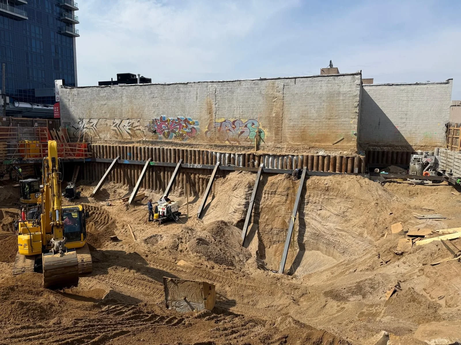 Construction site with excavation equipment and steel supports, with graffiti on a concrete wall in the background.